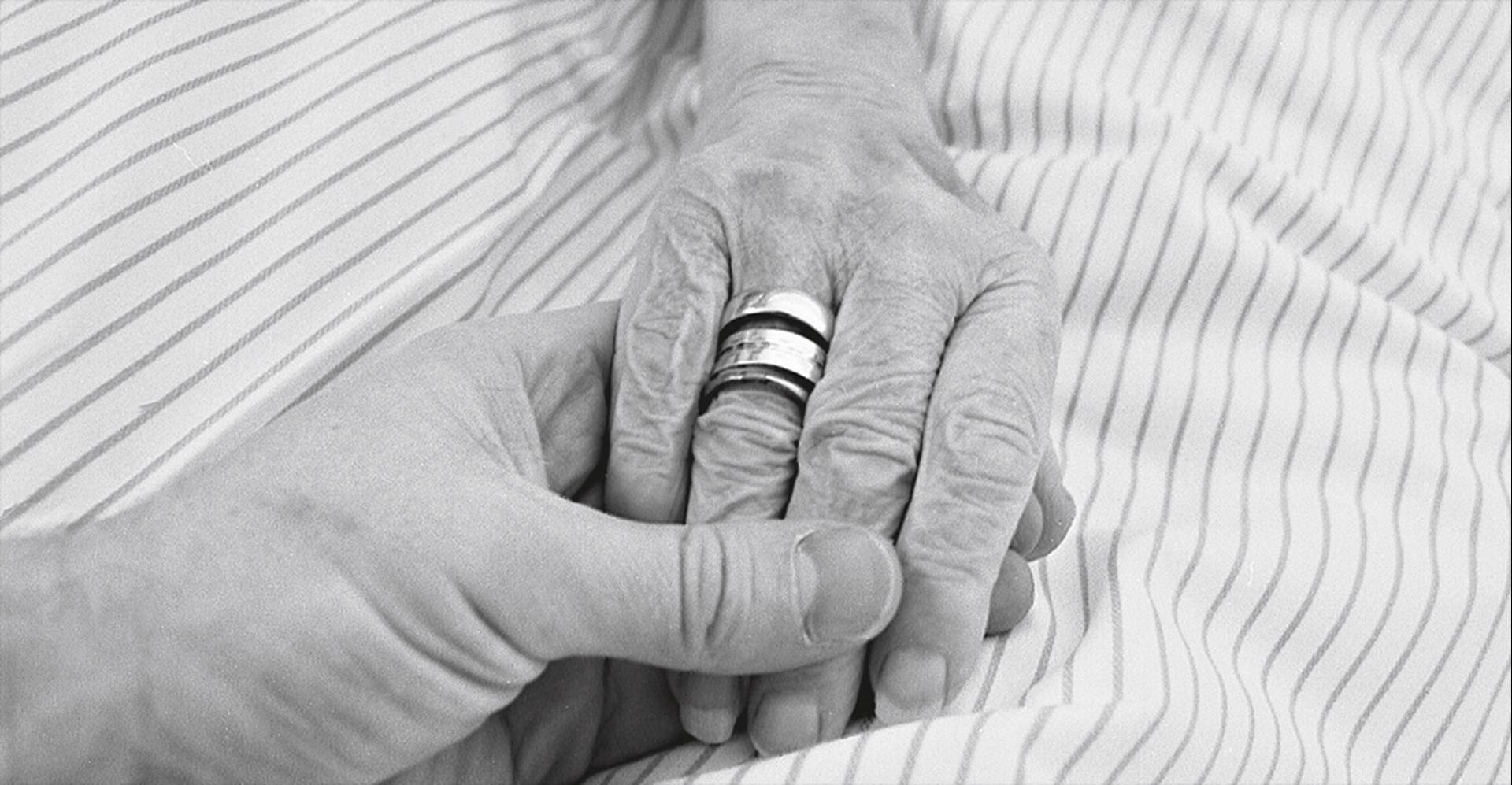 Die Hand einer Seniorin mit einigen Ringen am Ringfinger, die auf einer Bettdecke liegt und von einer anderen Hand gehalten wird (Foto in schwarz-weiß)