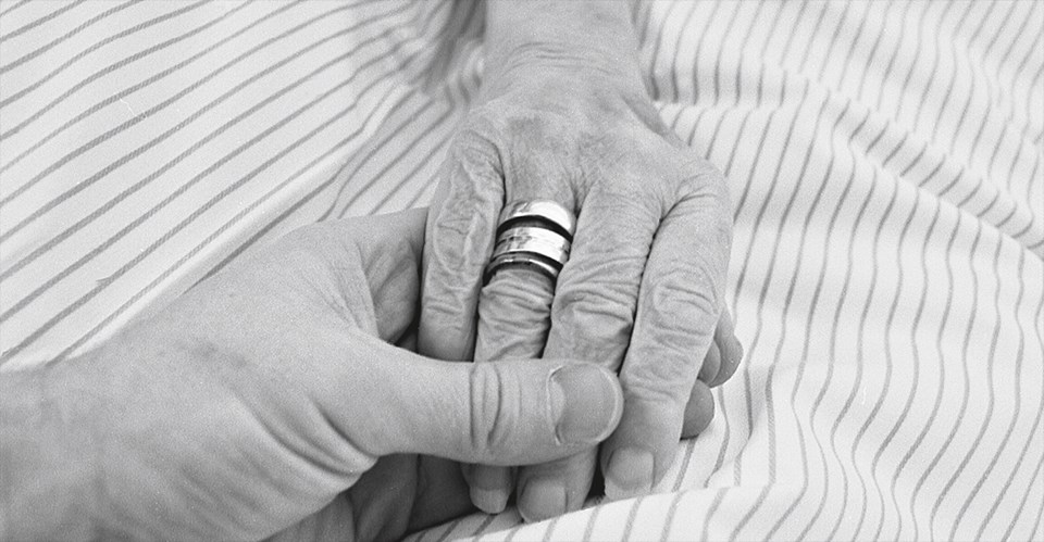 Die Hand einer Seniorin mit einigen Ringen am Ringfinger, die auf einer Bettdecke liegt und von einer anderen Hand gehalten wird (Foto in schwarz-weiß)