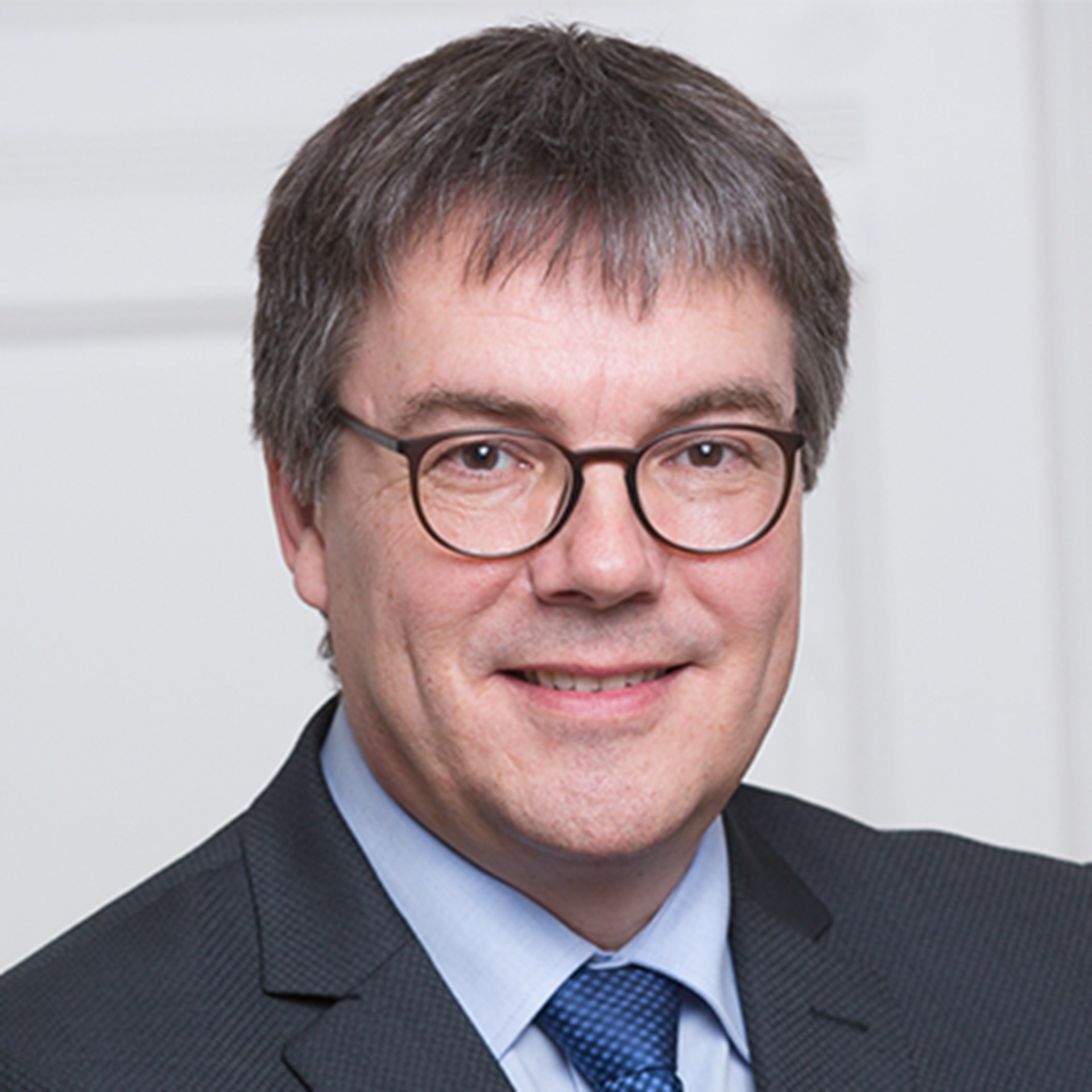 Lächelnder Mann mit grauen Haaren und Brille, der ein blaues Hemd, eine dunkelblaue Krawatte und eine schwarze Anzugjacke trägt