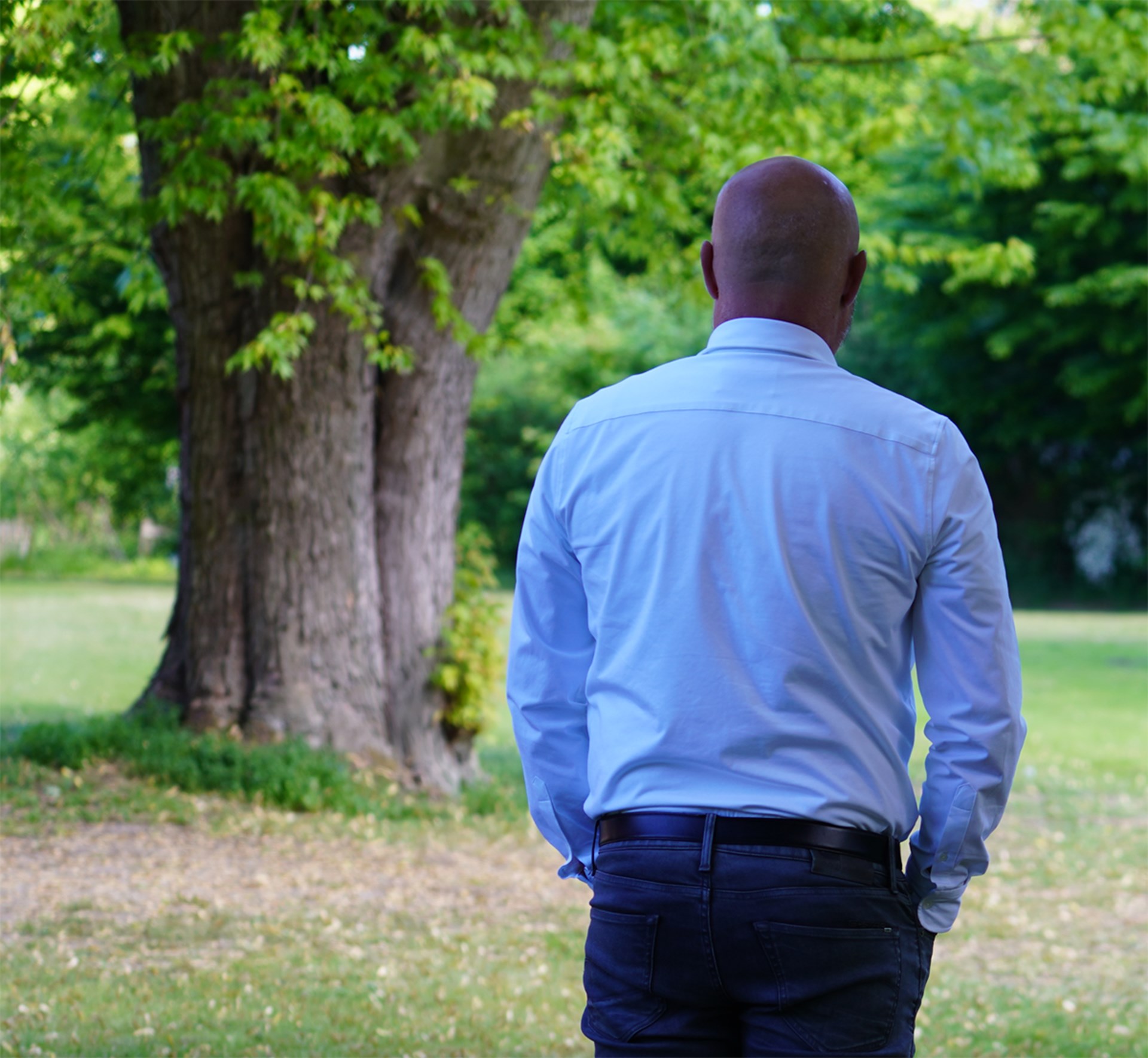 Ein schwarzer Mann mit Glatze, der ein weißes Hemd mit einer dunklen Jeans trägt und auf einer Wiese mit dem Rücken zur Kamera steht. In seiner Nähe steht ein Baum.