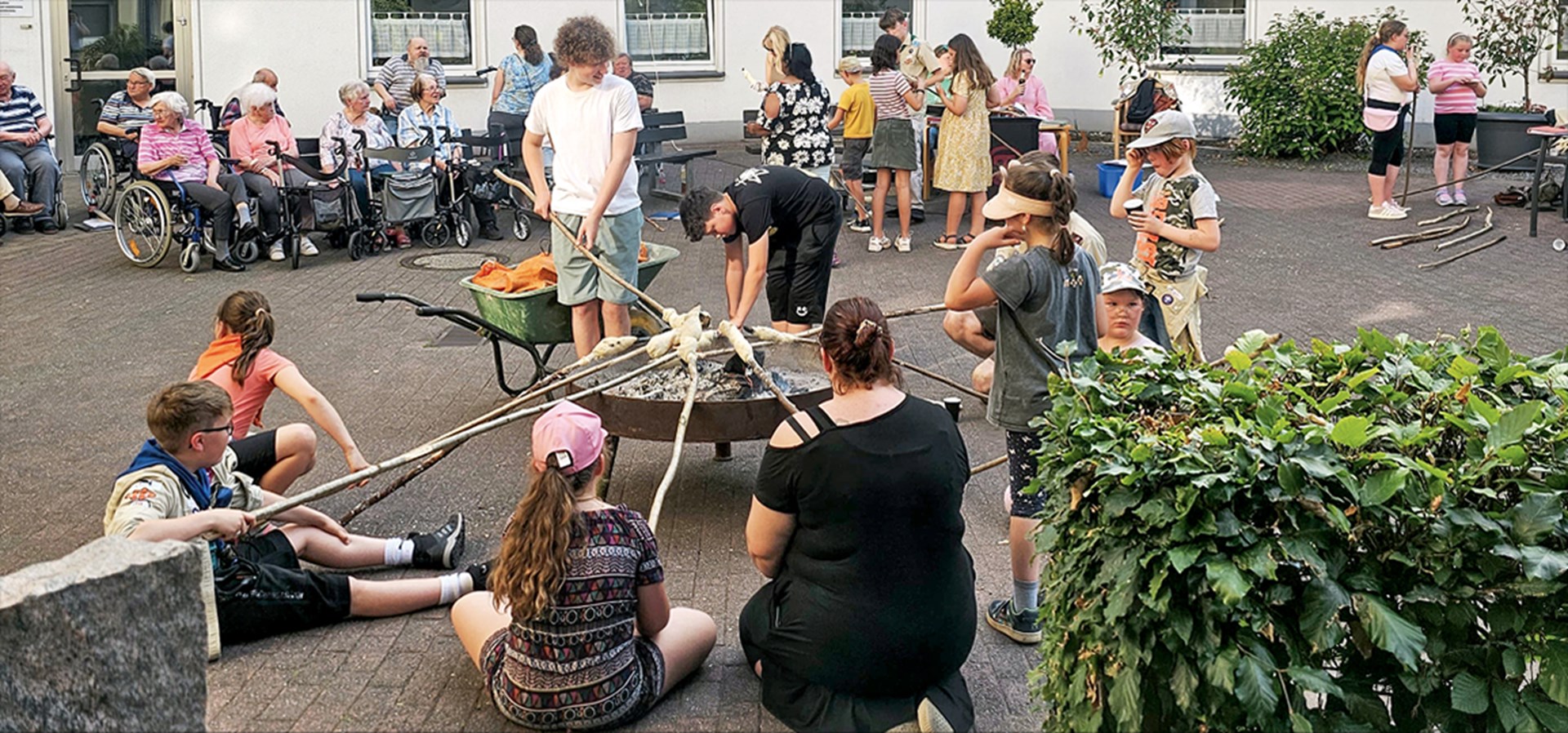 Eine Gruppe von Kinder und Jugendlichen sitzen bzw. stehen in einem Hinterhof um eine Feuerstelle und backen Stockbrot. Im Hintergrund sind weitere Menschen zu sehen, die in Gruppen zusammenstehen.