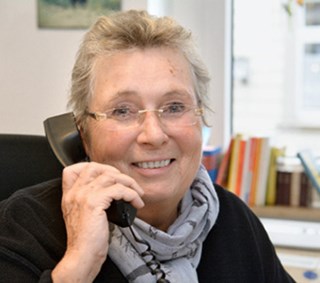 Lächelnde Frau mit grauen Haaren und Brille, die einen grauen Schal mit einer schwarzen Jacke trägt und in einem Büro an einem Schreibtisch sitzt. Sie hält einen Telefonhörer an ihr rechtes Ohr.
