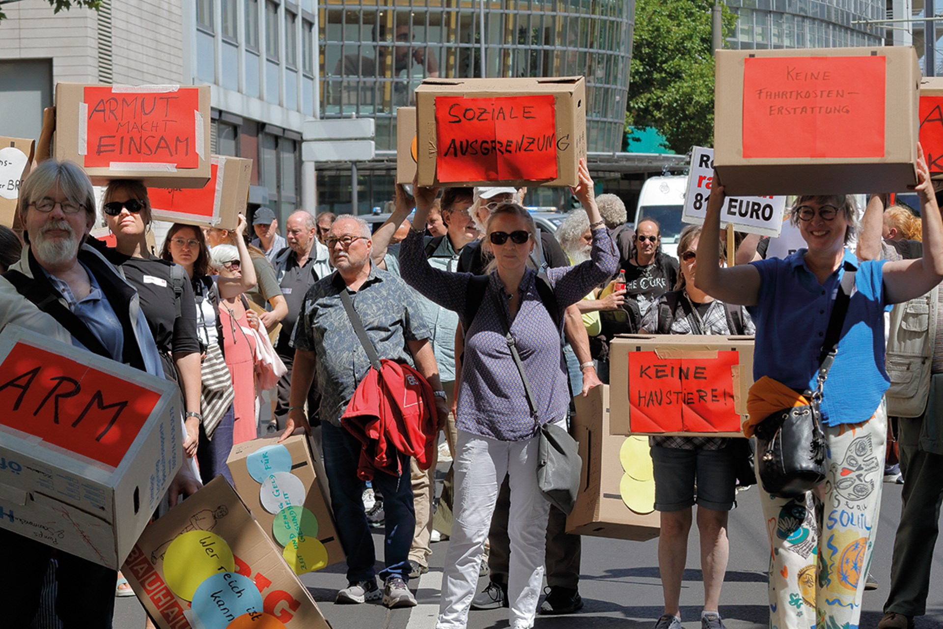 Eine Gruppe von Demonstrierenden, die mit Umzugskartons, auf denen rote Schilder mit Stichworten zum Thema 'Armut' kleben, bei einem sonnigen Tag durch die Kölner Innenstadt ziehen