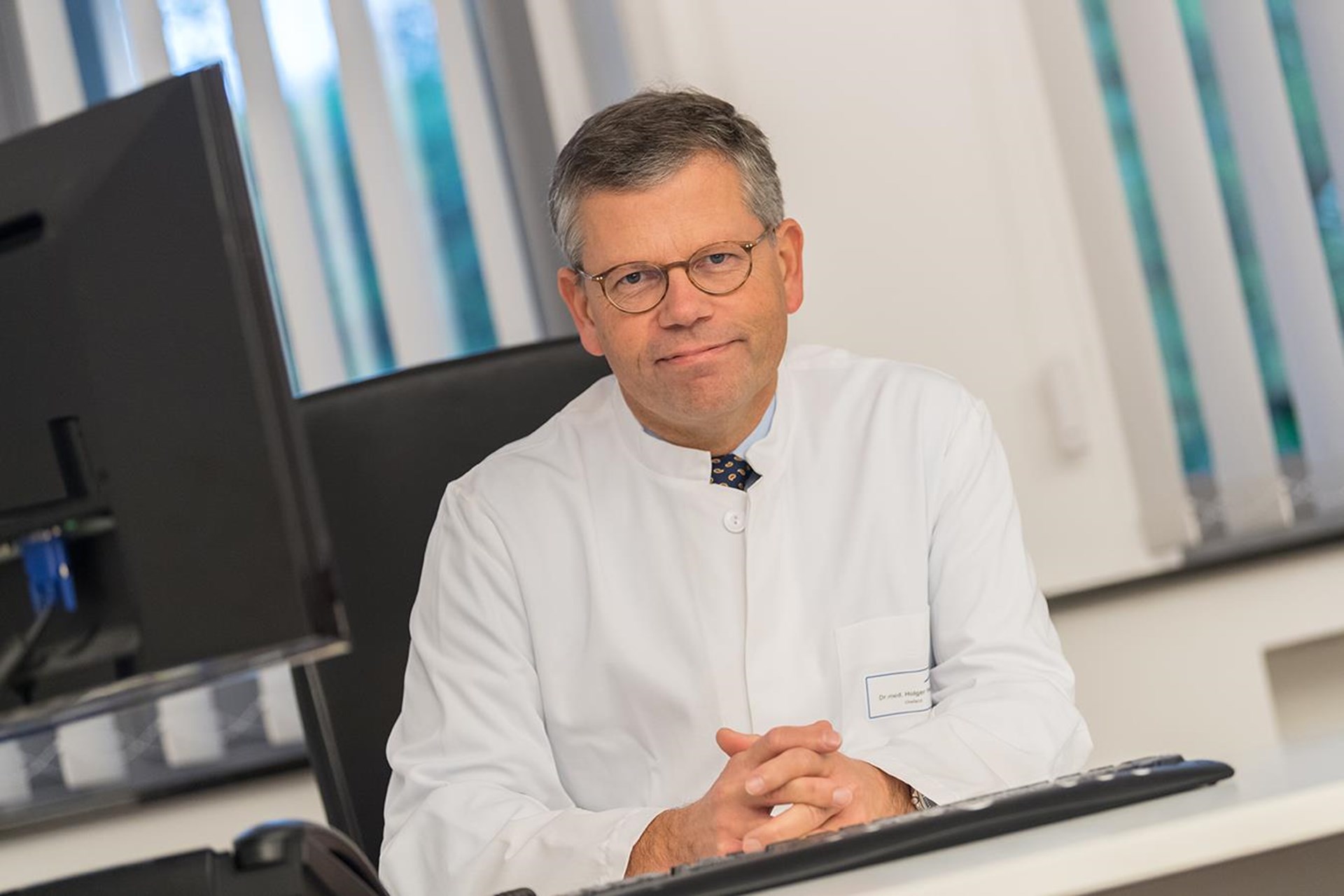 Ein Arzt sitzt an seinem Schreibtisch vor einem Computerbildschirm und blickt in die Kamera