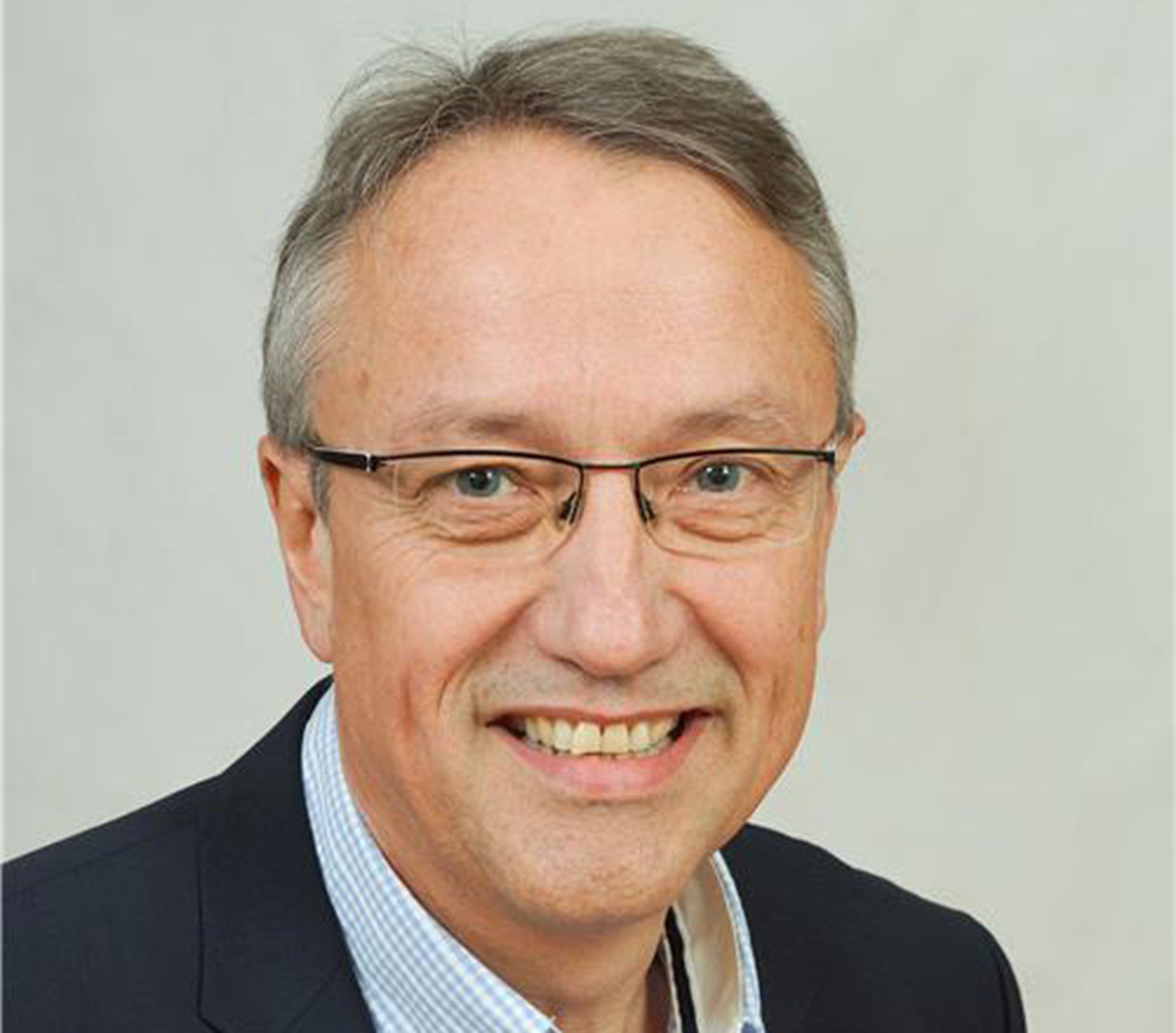 Lächelnder Mann mit grauen Haaren und Brille, der ein blaues Hemd und eine dunkelblaue Anzugjacke trägt