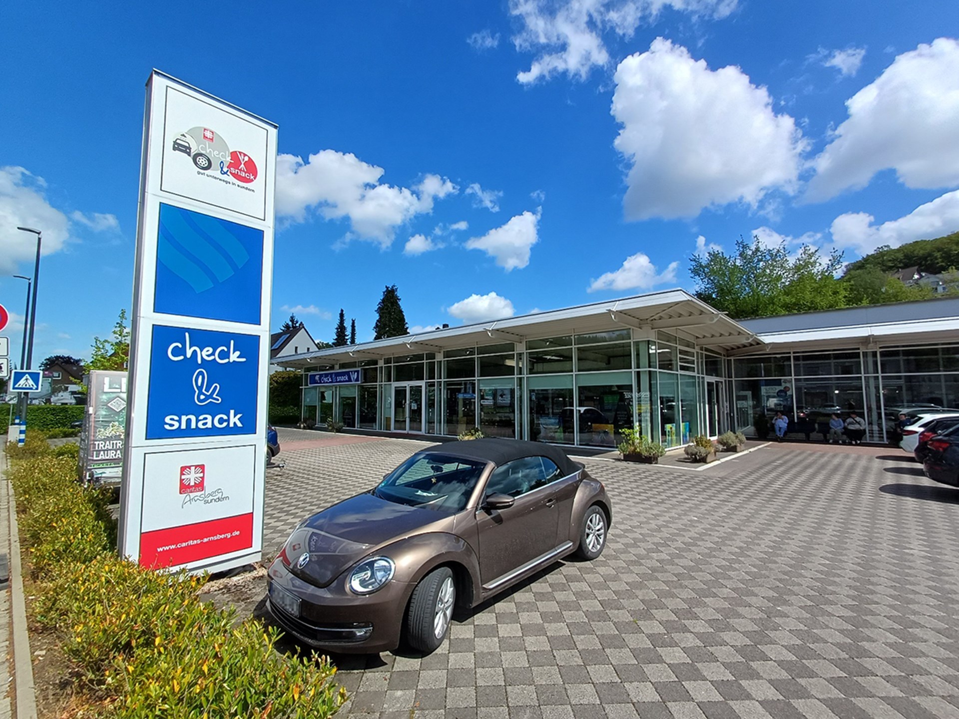 Der gepflasterte Vorplatz des Gebäudes von 'check & snack' der Caritas Arnsberg-Sundern an einem sonnigen Tag. Im Vordergrund ist ein großes Hinweisschild und parkender VW Beetle zusehen.
