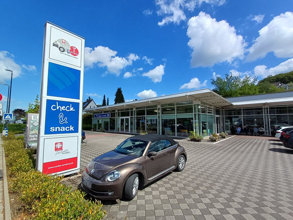 Der gepflasterte Vorplatz des Gebäudes von 'check & snack' der Caritas Arnsberg-Sundern an einem sonnigen Tag. Im Vordergrund ist ein großes Hinweisschild und parkender VW Beetle zusehen.