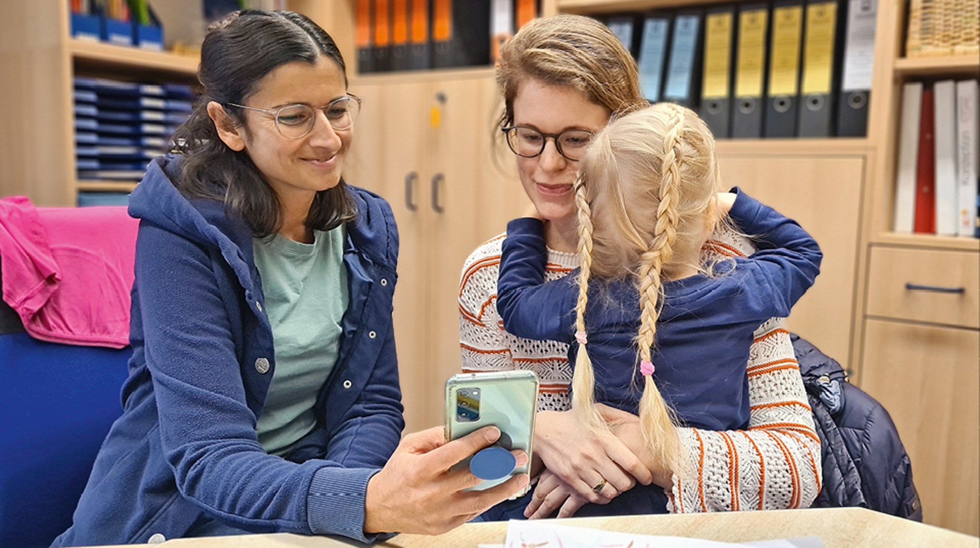 Eine Kita-Leiterin und eine Mutter sitzen in einem Büro zusammen und blicken auf ein Handy. Die Mutter hat dabei ihr Mädchen auf dem Arm.