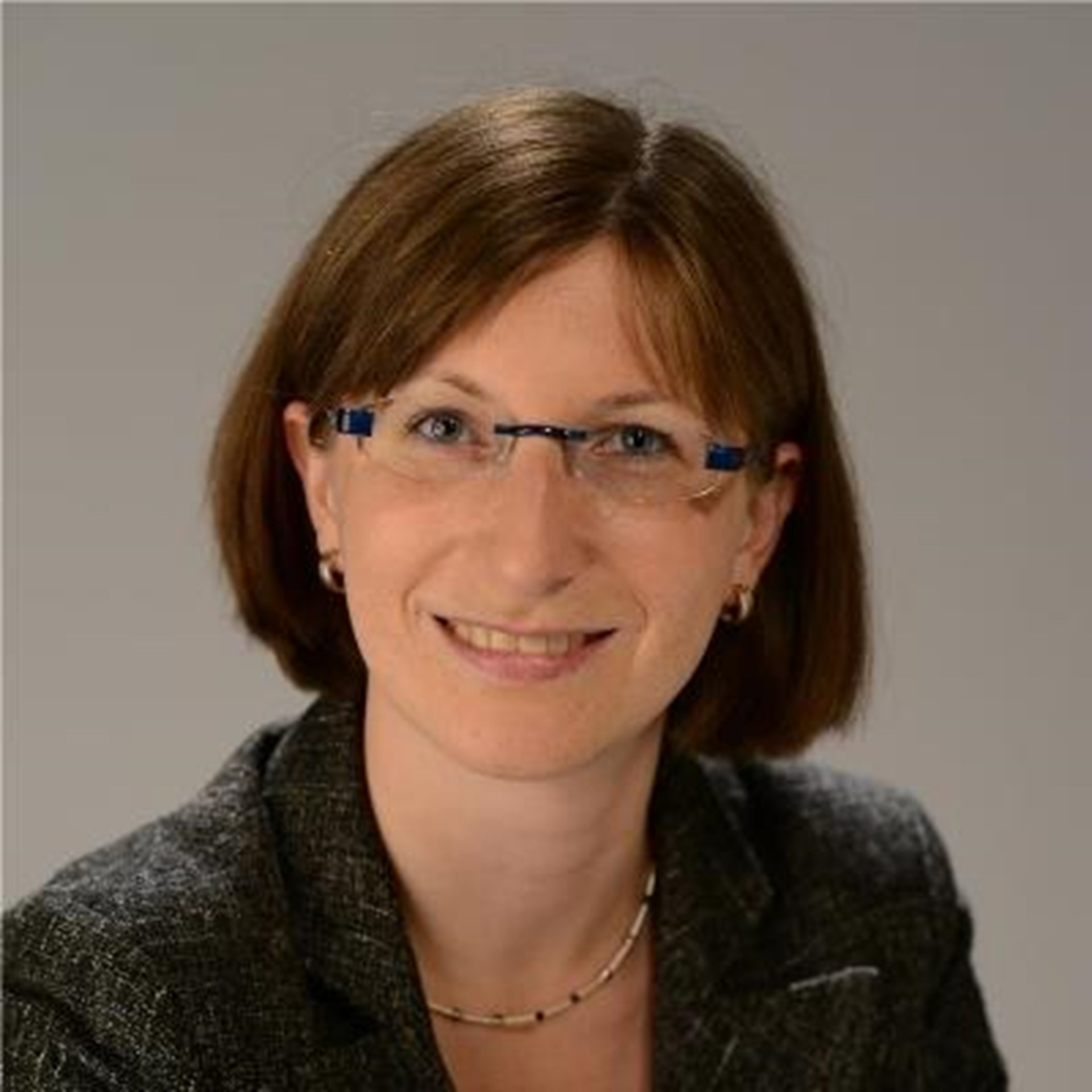 Lächelnde Frau mit schulterlangen, braunen Haaren und Brille, die eine dunkelgraue Anzugjacke trägt und ihre Arme verschränkt.