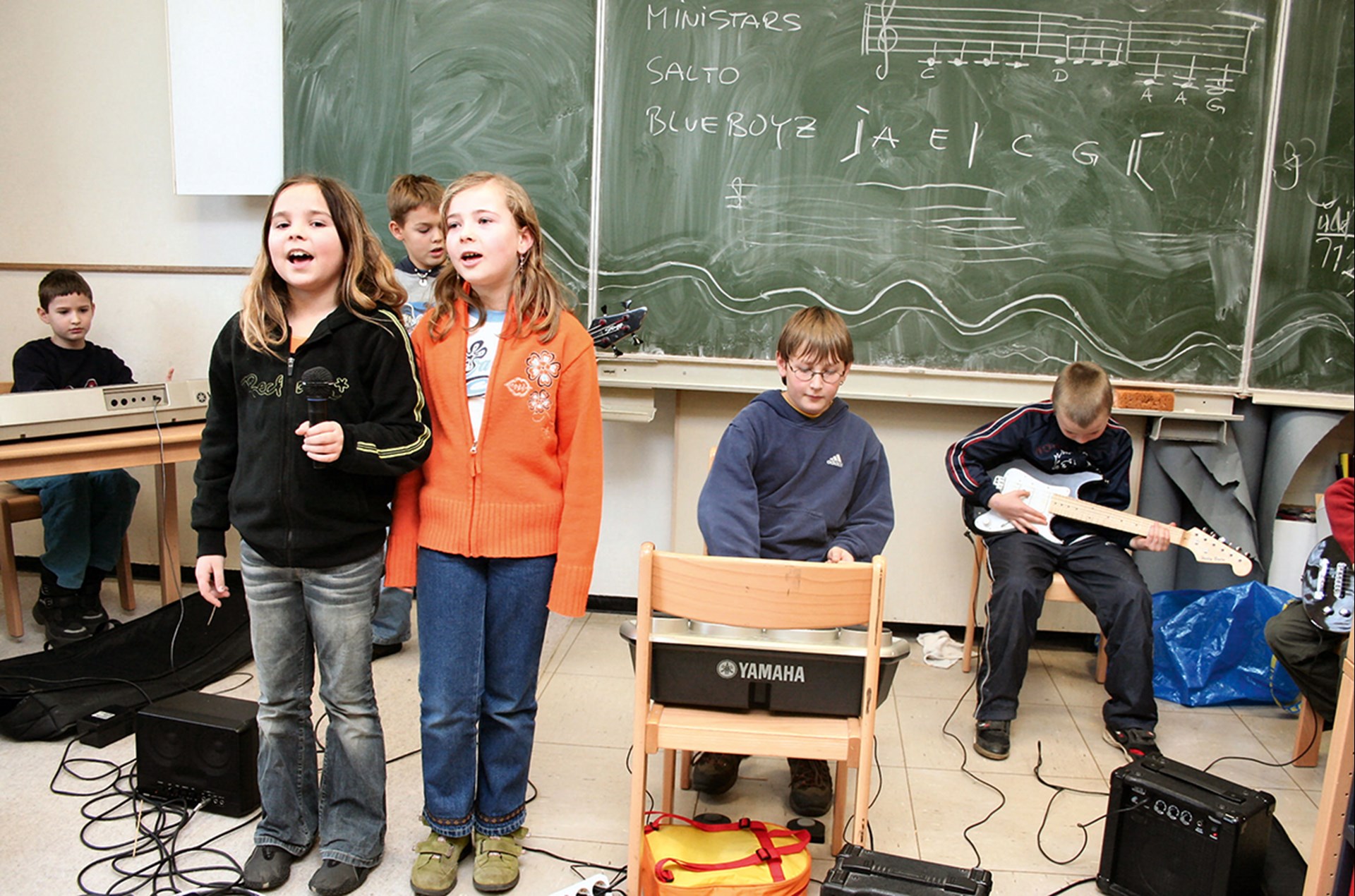 Drei Schüler befinden sich mit verschiedenen Musikinstrumenten in einem Klassenzimmer einer OGS der Hubertusschule in Kevelaer und mu­si­zie­ren. Davor stehen zwei Mädchen mit einem Mikrofon und singen.