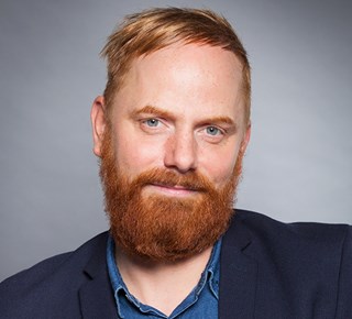 Mann mit orangen Haaren und Vollbart, der ein blaues Hemd und eine blaue Anzugjacke trägt