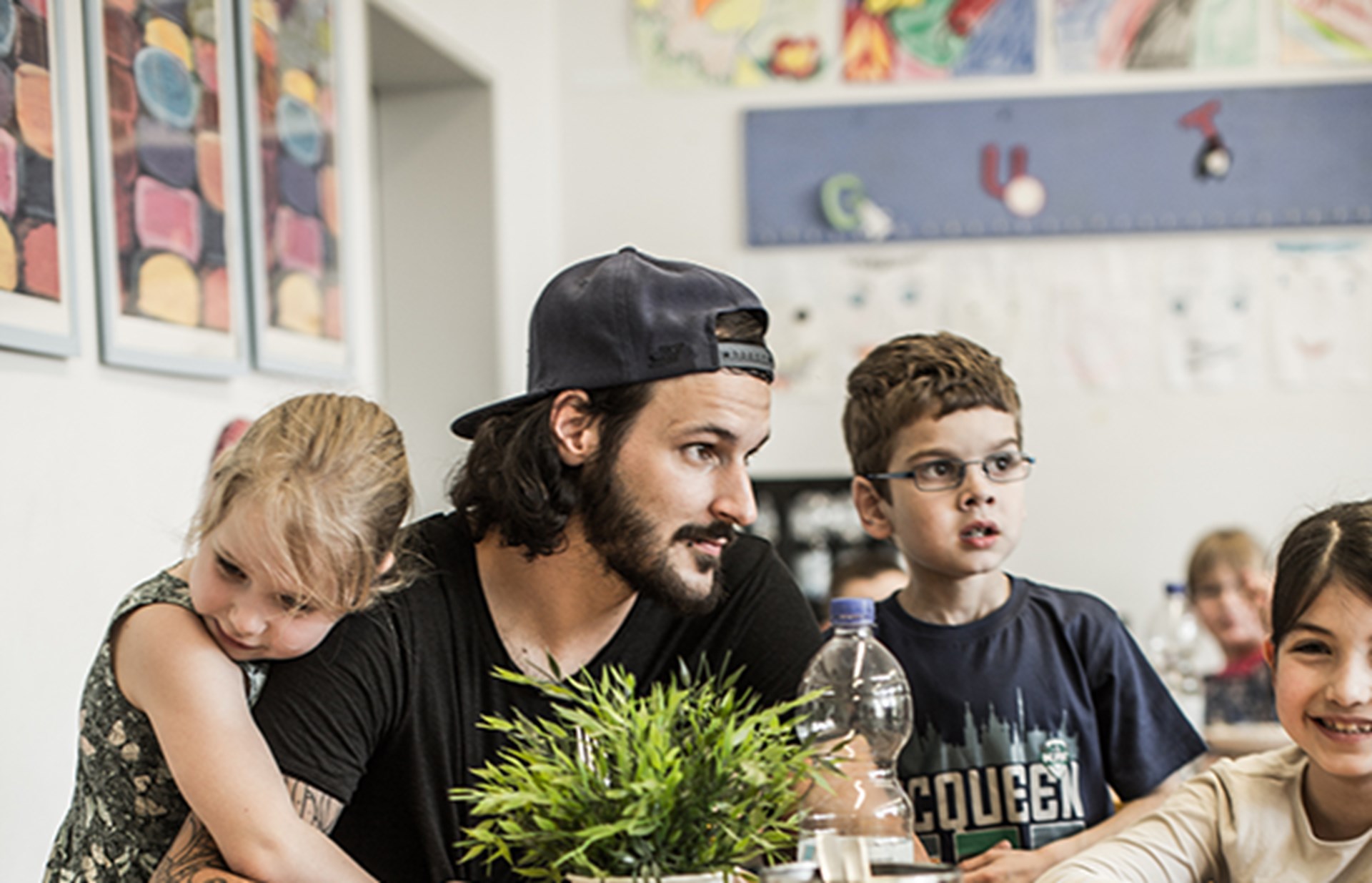 Ein junger Betreuer in einer OGS sitzt mit einigen Kindern an einem Tisch. Ein Mädchen umarmt ihn von hinten. Ein Junge beugt seinen Kopf nachhinten und blickt in die Kamera.