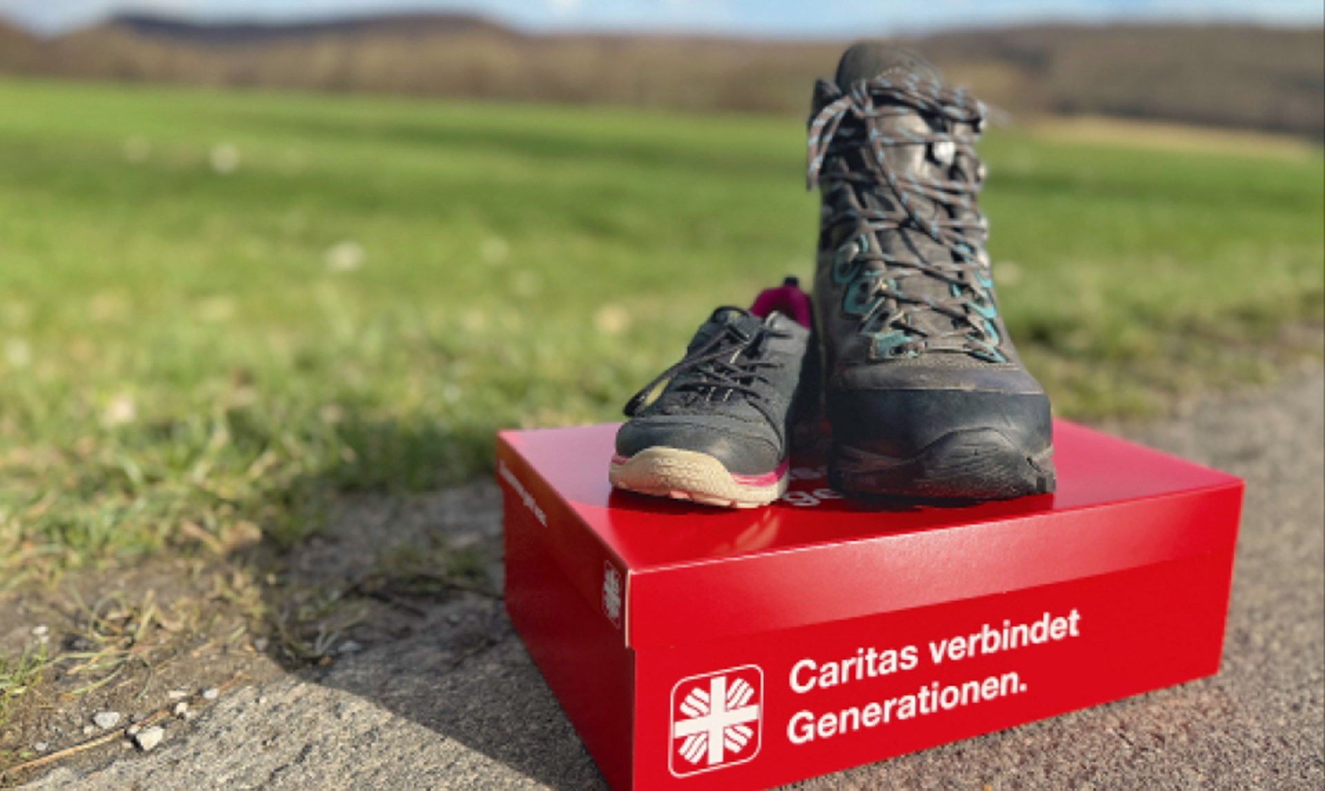 Ein roter Caritas-Schuhkarton mit der Aufschrift 'Caritas verbindet Generationen.' der am Rande eines Wanderweges vor einer Wiese steht. Auf dem Karton stehen ein kleiner und größerer Wanderschuh.