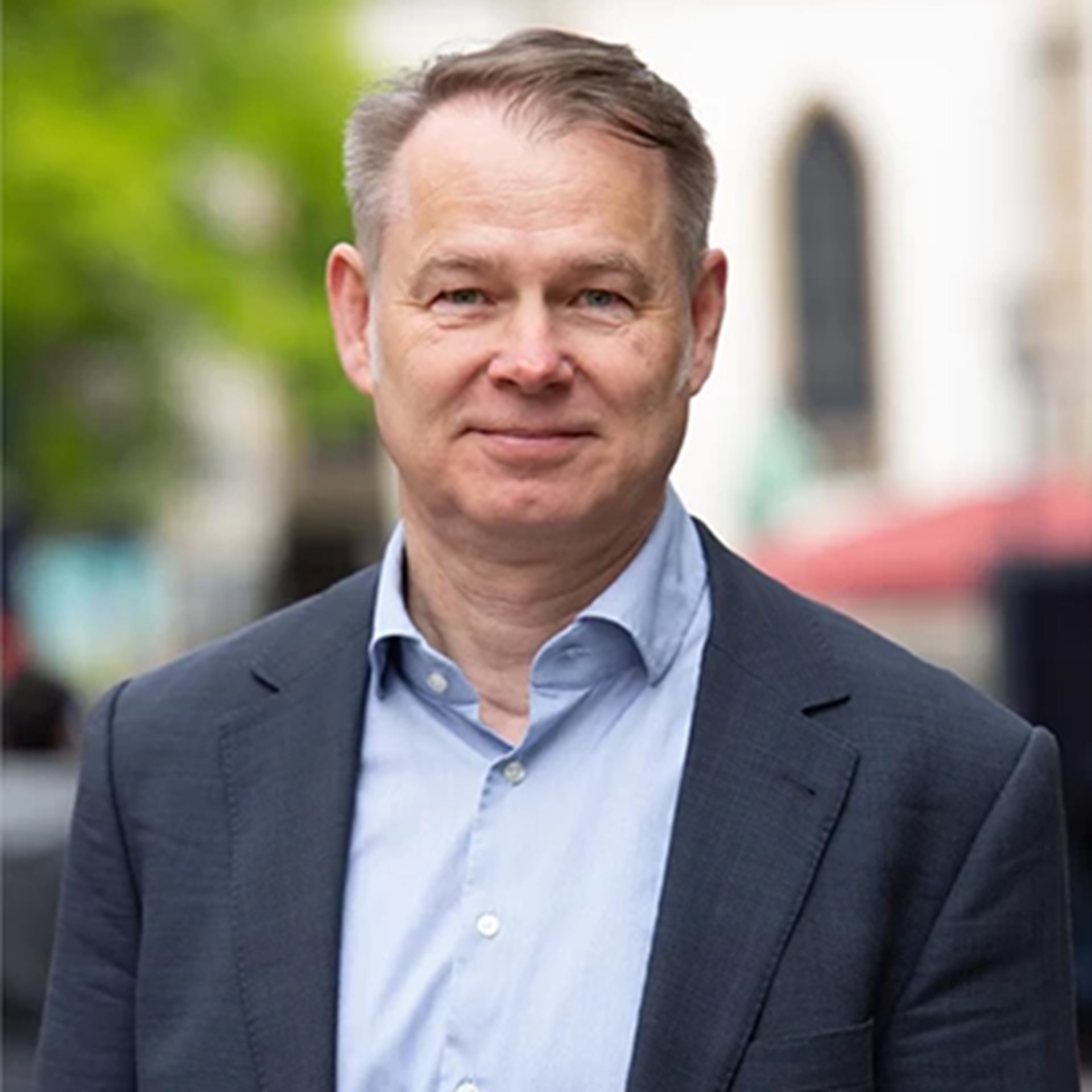 Lächelnder Mann mit blonden Haaren, der ein hellblaues Hemd und eine dunkle Anzugjacke trägt. Er steht in der Fußgängerzone der Essener Innenstadt.