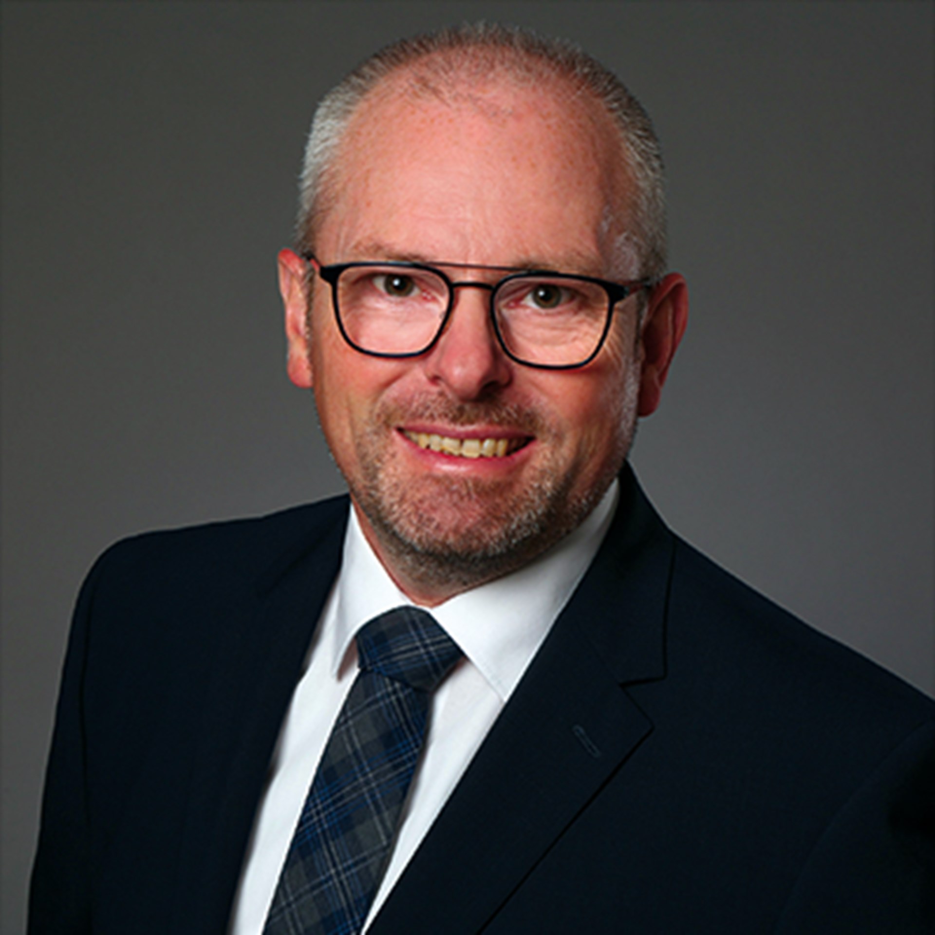 Lächelnder Mann mit grauen Haaren, Brille und Bart, der ein weißes Hemd, eine dunkle Krawatte und eine schwarze Anzugjacke trägt