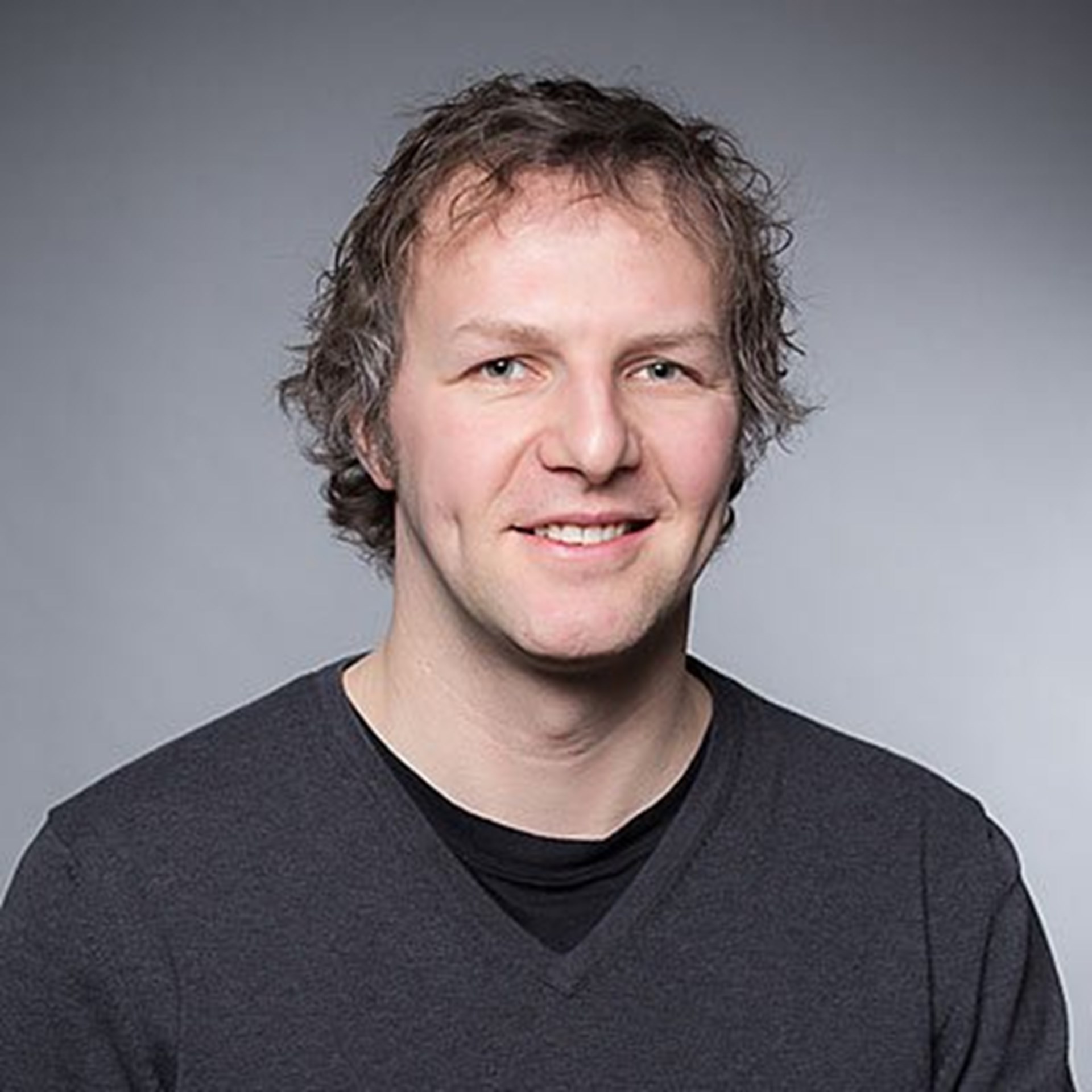 Lächelnder Mann mit blond-grauen Haaren, der ein schwarzes T-Shirt mit einem dunkelgrauen Pullover trägt