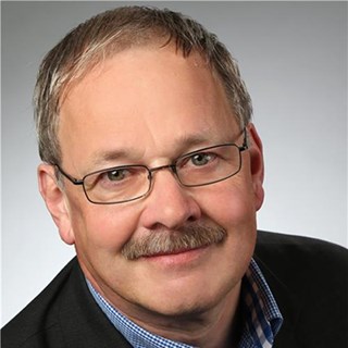 Mann mit kurzen, blonden Haaren, Schnäuzer und Brille, der ein weiß-blau-kariertes Hemd und eine schwarze Anzugjacke trägt
