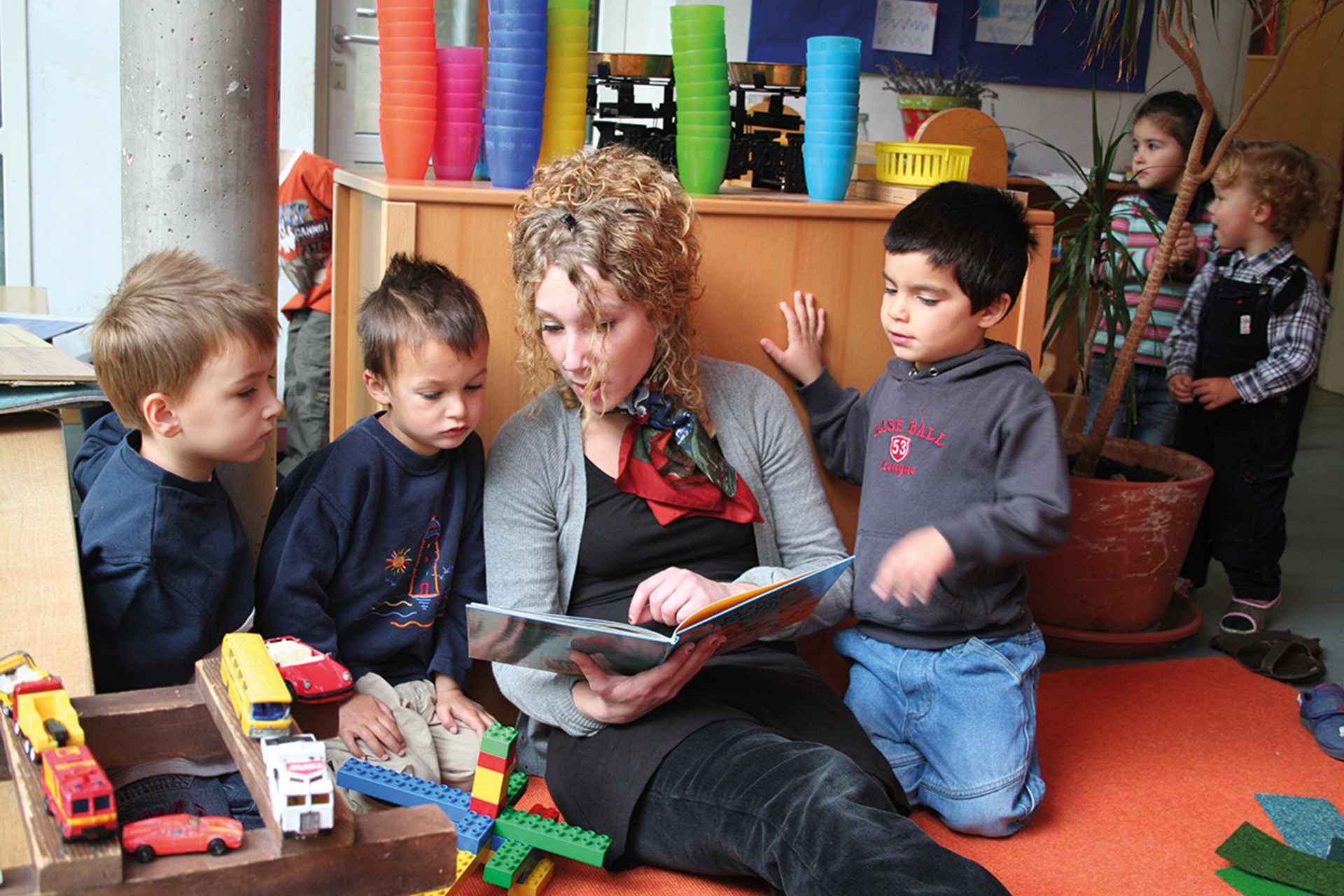 Eine Erzieherin sitzt in einer Kindertageseinrichtung an einen Schrank gelehnt auf dem Boden und liest in einem Kinderbuch. Drei Jungen knien neben ihr und blicken mit in das Kinderbuch. Im Hintergrund sind spielende Kinder zu sehen.