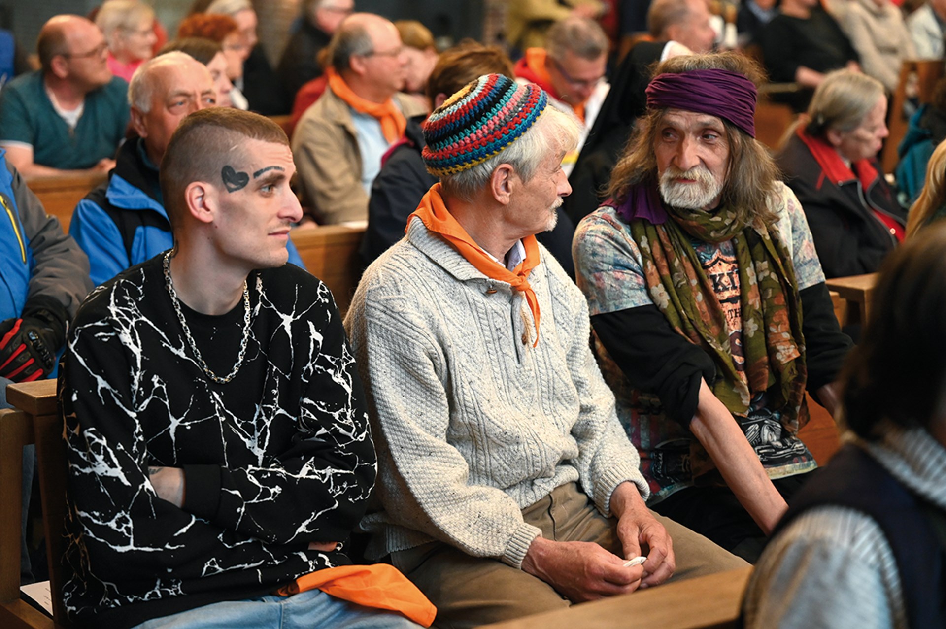 Drei Obdachlose, die in einer gut gefüllten Kirche auf einer Kirchenbank sitzen. Zwei der Obdachlosen schauen sich gegenseitig an.
