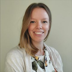 Lächelnde, junge Frau mit langen, blonden Haaren, die ein Hemd mit Pflanzenmuster und eine helle Jacke trägt