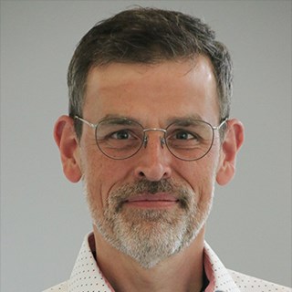 Sanft lächelnder Mann mit grauen Haaren, 3-Tage-Bart und Brille, der ein weißes Hemd trägt