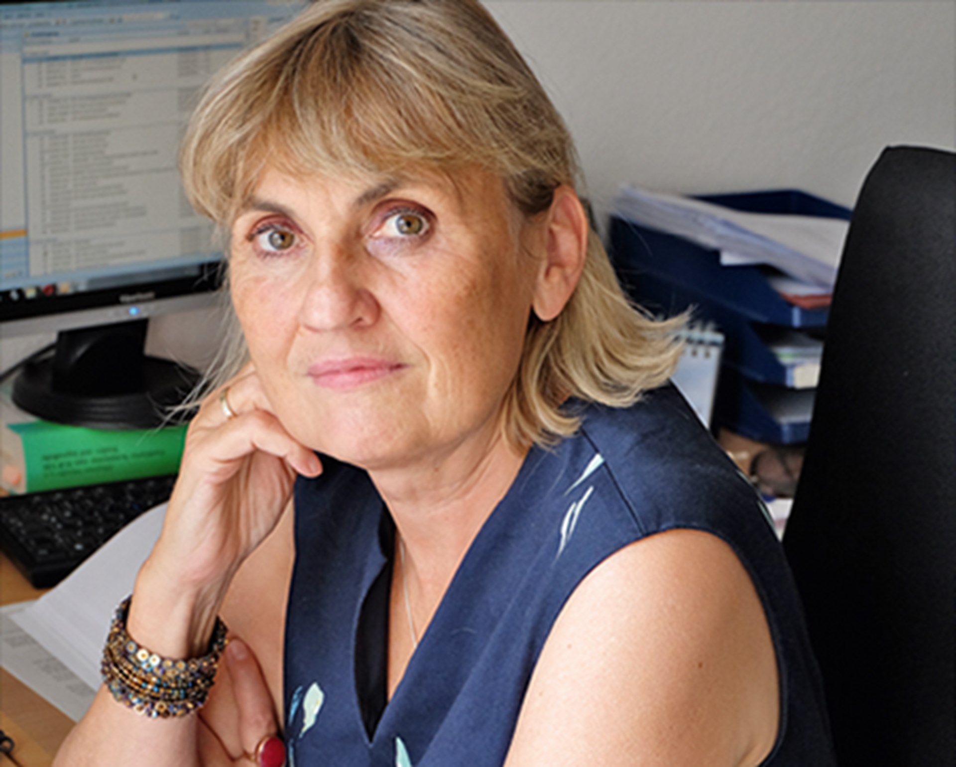 Frau mit schulterlangen, blonden Haaren, die ein ärmelloses, blaues Shirt trägt und in einem Büro an einem Schreibtisch sitzt. Sie stützt ihren Kopf auf ihrem rechten Arm ab.