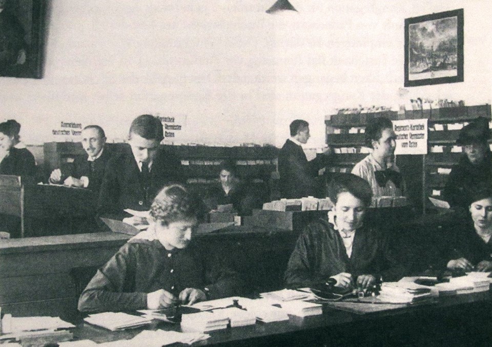 Historische Aufnahme in Schwarz-Weiß: Einige weibliche und männliche Mitarbeiter stehen bzw. sitzen in einem Büroraum mit Schreibbänken und Regalen und arbeiten