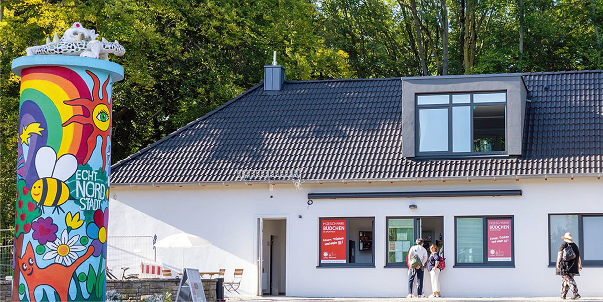 Außenansicht des Caritas-Büdchens im Dortmunder Hoeschpark, neben dem eine bunt bemalte Litfaßsäule steht. Vor dem Büdchen stehen drei Personen.