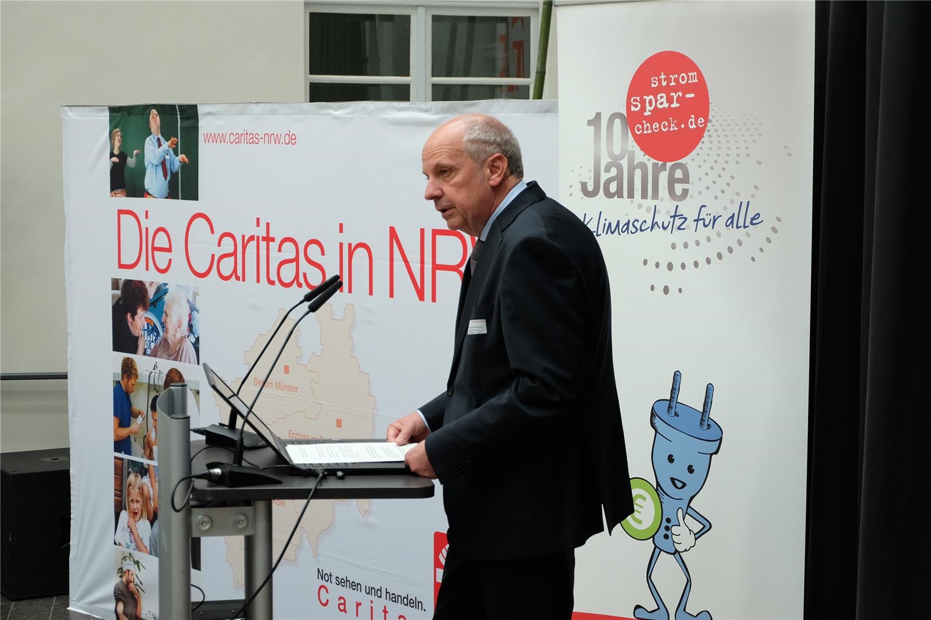 Ein Mann steht an einem Rednerpult. Im Hintergrund ist ein Faltdisplay der Caritas in NRW zu sehen.