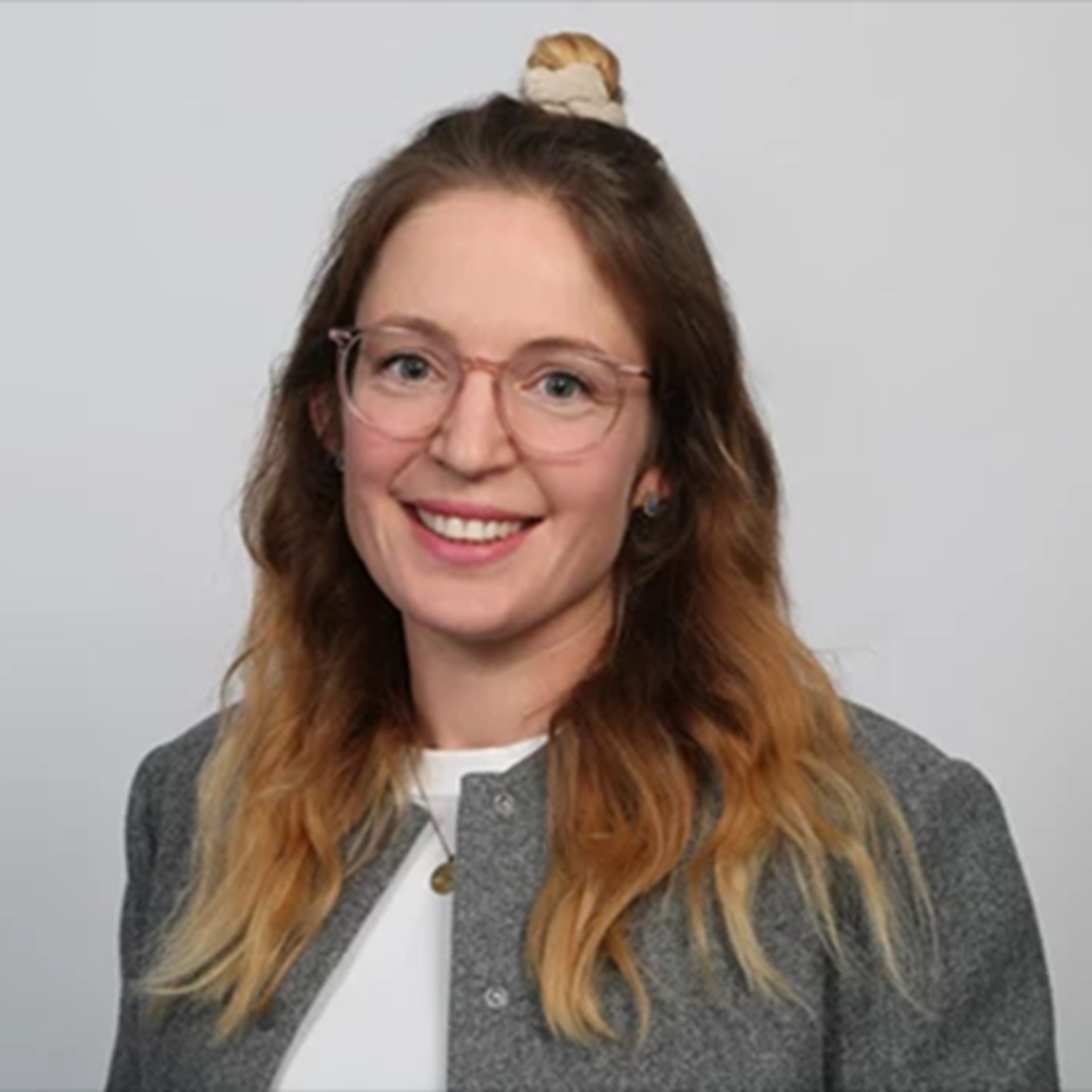 Lächelnde Frau mit langen, blonden Haaren und Brille, die ein weißes Shirt und eine graue Jacke trägt.