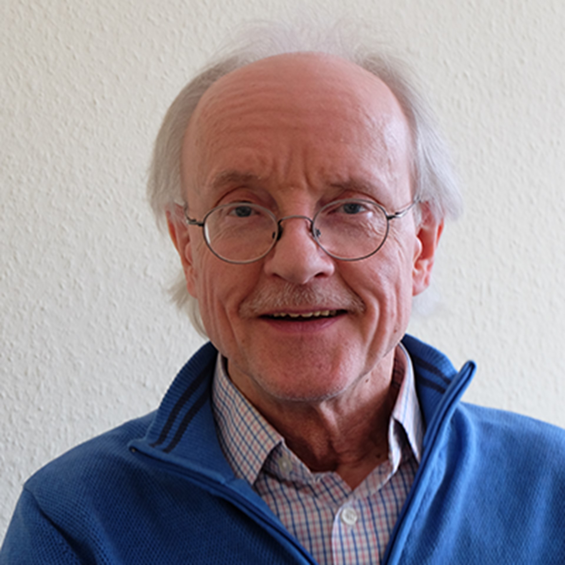 Sanft lächelnder Mann mit grauem Haar und Halbglatze, der eine Brille und ein helles Hemd mit einer blauen Jacke trägt