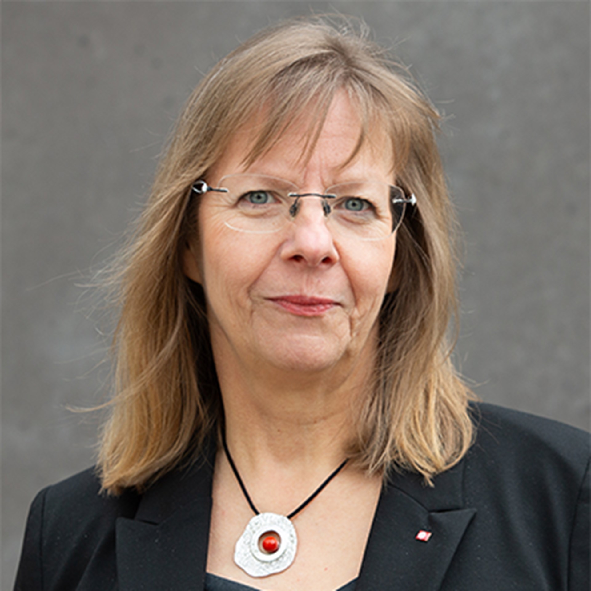 Frau mit schulterlangen, blonden Haaren und Brille, die einen schwarzen Anzug und einer Halskette mit einem silbernen Anhänger und einem roten Stein trägt