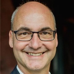 Lächelnder Mann mit grauen Haaren, Halbglatze und Brille, der ein weißes Hemd und eine graue Anzugjacke trägt