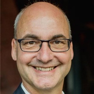 Lächelnder Mann mit grauen Haaren, Halbglatze und Brille, der ein weißes Hemd und eine graue Anzugjacke trägt