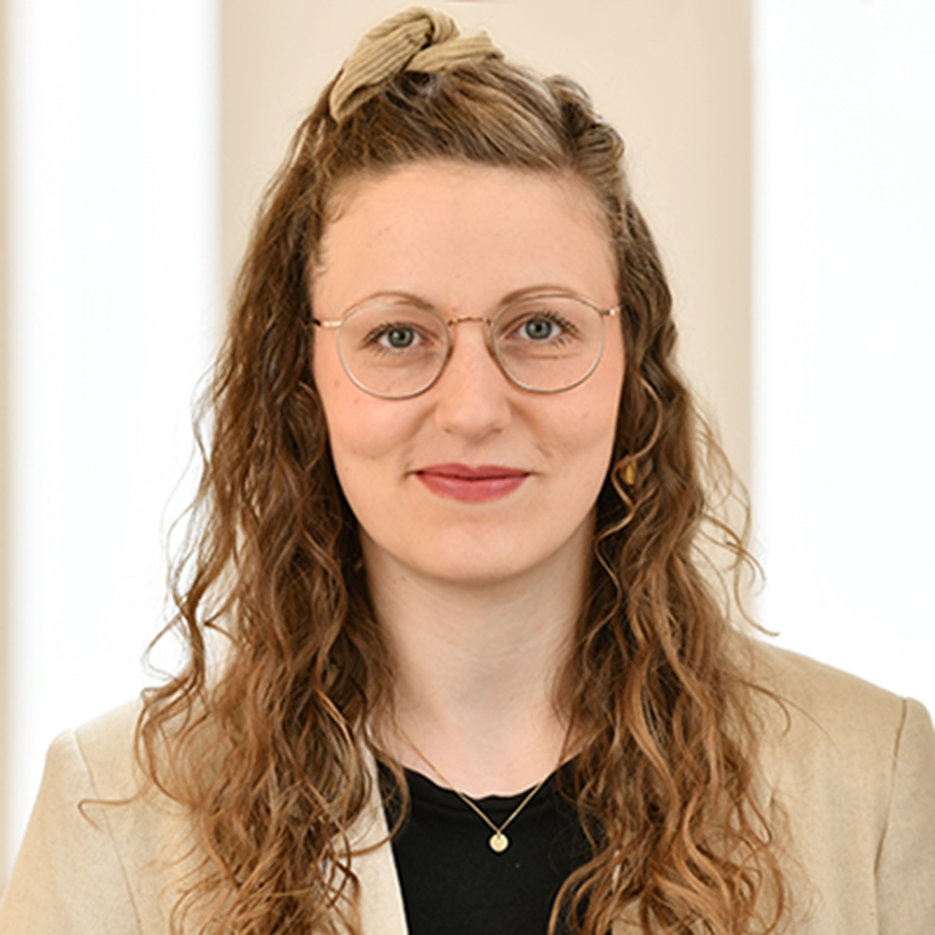 Lächelnde Frau mit langen, blonden Haaren und Brille, die ein schwarzes Shirt und eine helle Anzugjacke trägt.