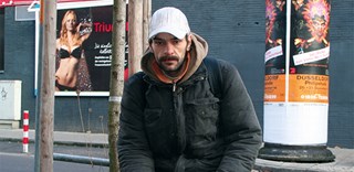 Ein obdachloser Mann mit einer dicken Jacke und einer weißen Kappe sitzt am Straßenrand auf einer Metall-Begrenzung vor einer Grünfläche und blickt in die Kamera. Er hält eine weiße Caritas-Respekttüte in der Hand. Im Hintergrund sind eine Werbetafel und eine Litfaßsäule zu sehen.