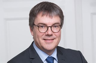 Lächelnder Mann mit grauen Haaren und Brille, der ein blaues Hemd, eine dunkelblaue Krawatte und eine schwarze Anzugjacke trägt. Er verschränkt die Arme.
