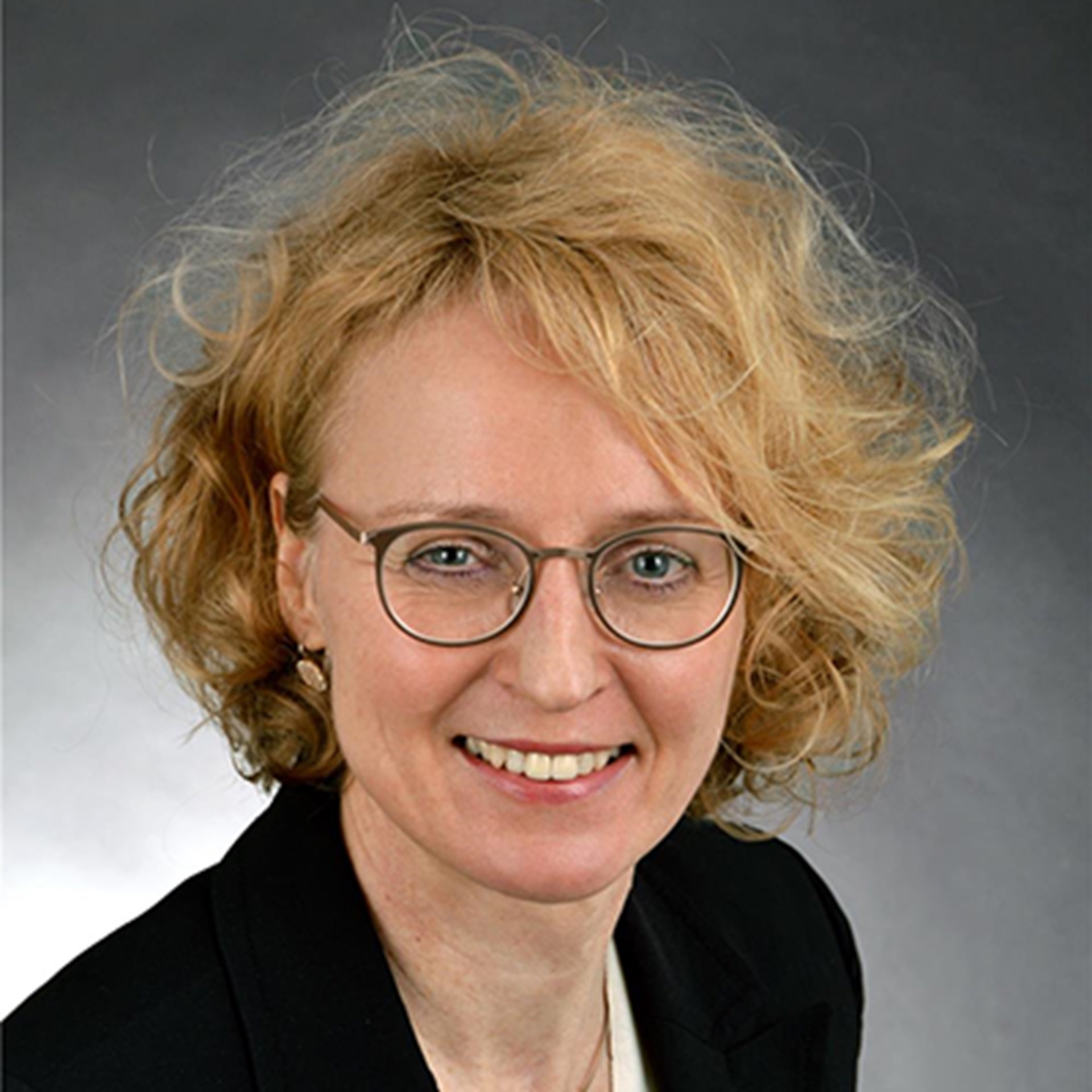 Lächelnde Frau mit blonden Haaren und Brille, die ein weißes Shirt mit einer schwarzes Anzugjacke trägt