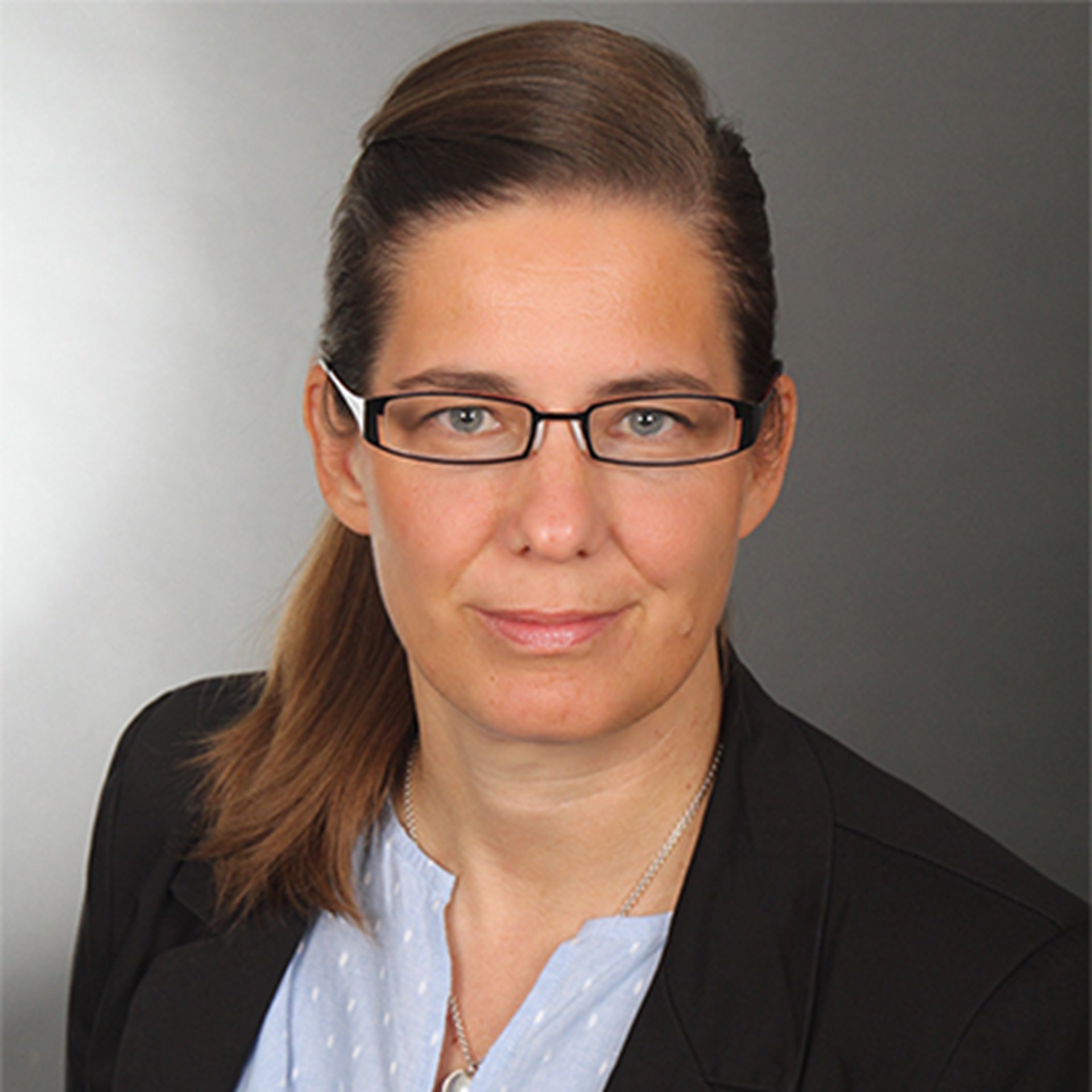 Sanft lächelnde Frau mit langen braunen Haaren und Brille, die ein hellviolettes Shirt und eine schwarze Anzugjacke trägt