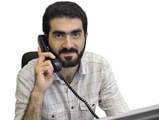 Lächelnder Mann mit schwarzen Haaren und Bart, der ein hellgrau-kariertes Hemd trägt und vor einem Computer sitzt. Er hält einen Telefonhörer an sein Ohr.