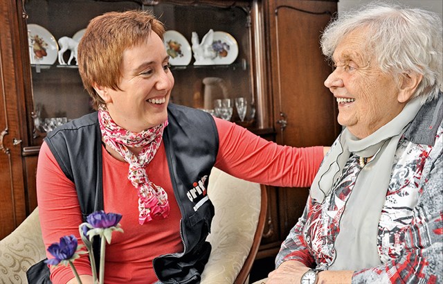 Eine seelsorgliche Begleiterin der Caritas sitzt mit einer Seniorin in einem Wohnraum und hält Ihre linke Hand an die Schulter der Seniorin. Beide lächeln sich an.