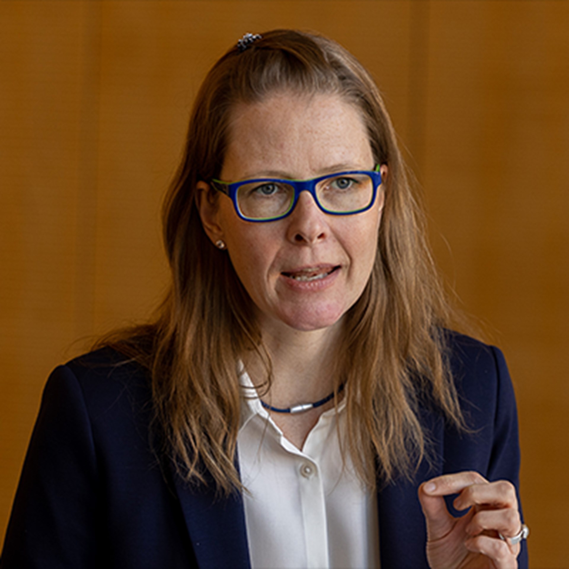 Sprechende Frau mit langen, braunen Haaren und blauer Brille, die ein weißes Hemd und eine schwarze Anzugjacke trägt und vor einer mit Holz verkleideten Wand sitzt