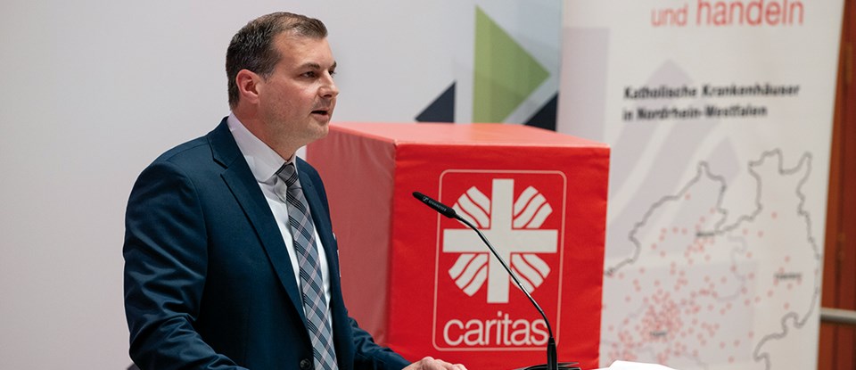 Ein Diözesan-Caritasdirektor steht beim 2. Kath. Krankenhaustag in Essen an einem Pult und hält eine Rede. Im Hintergrund ist ein roter Caritas-Würfel und ein Roll-up zu sehen.