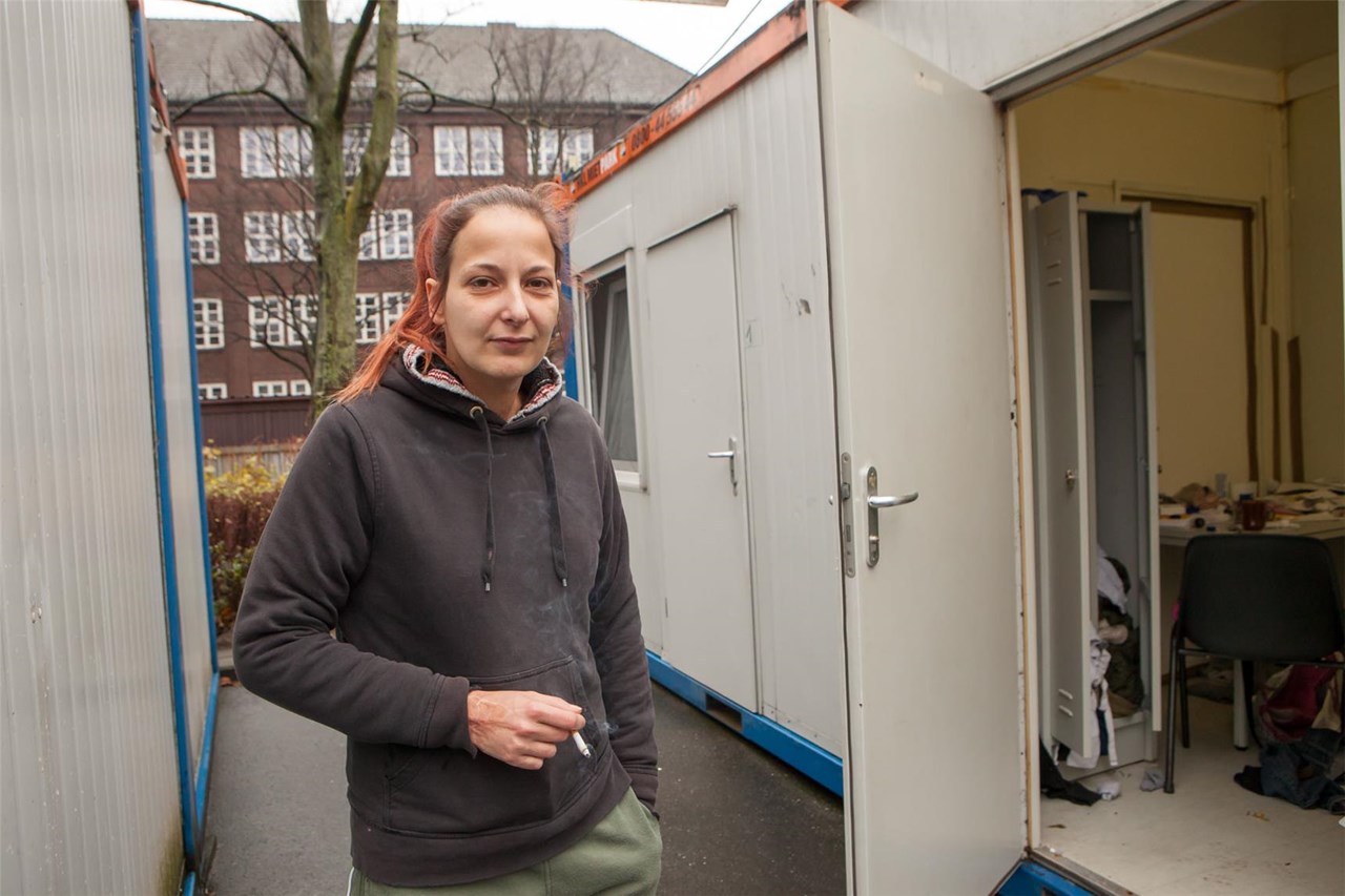 Frau mit Pferdeschwanz und Zigarette in der Hand neben Eingang zu ihrem Wohncontainer