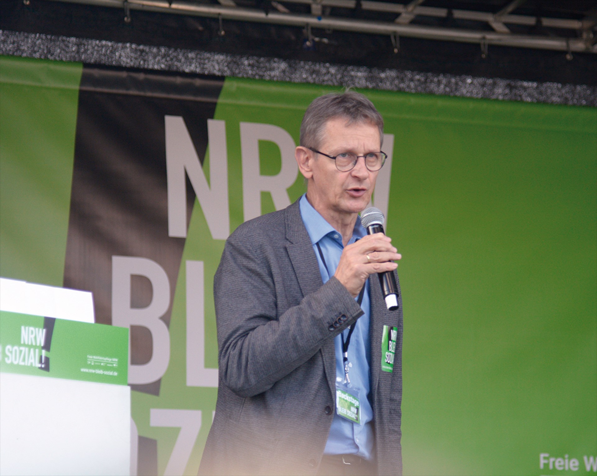 Ein Mann steht auf einer Bühne und redet in ein Handmikrofon. Im Hintergrund ist das Logo der Kampagne 'NRW bleib sozial!' auf grünem Hintergrund zu sehen.