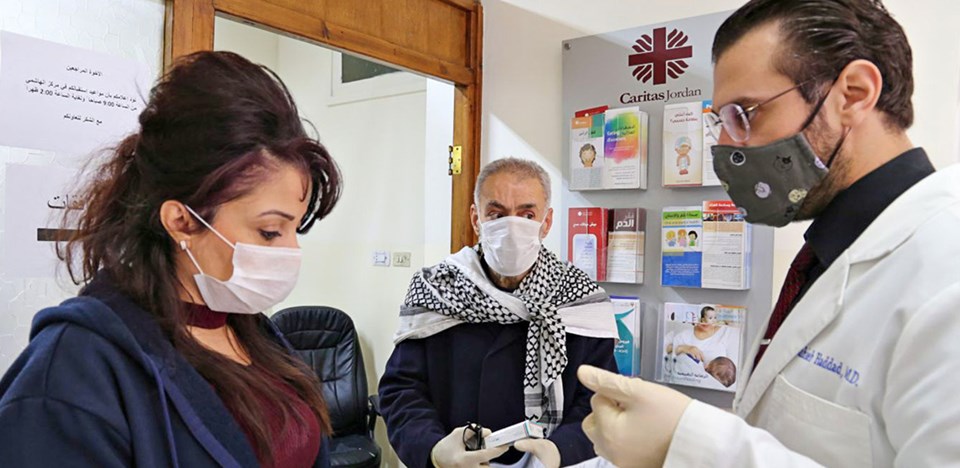 Ein Arzt spricht in den Räumlichkeiten der Caritas Jordanien mit einer Frau und einem Senior. Alle tragen Einweghandschuhe und Schutzmasken.