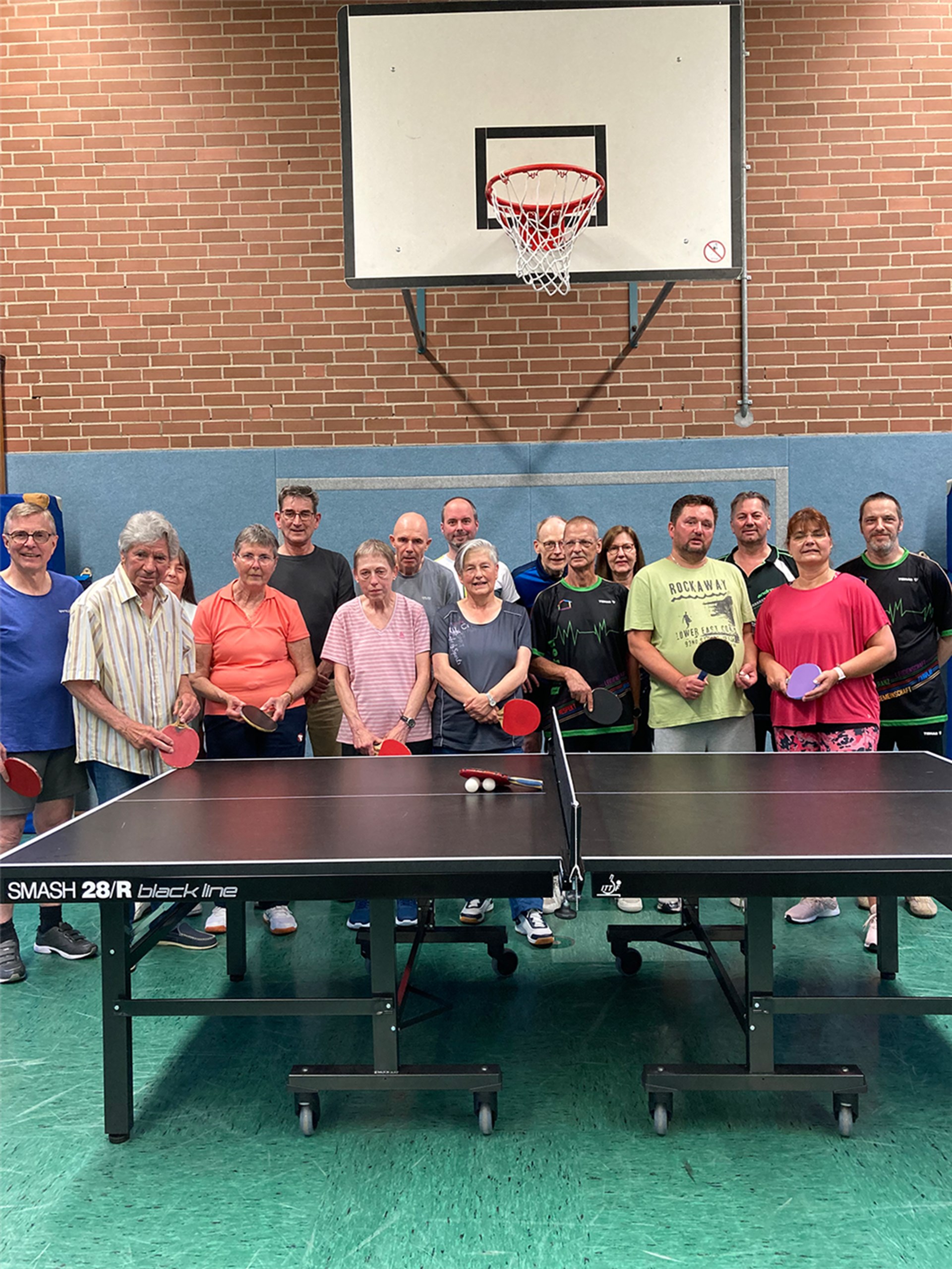 Ein Gruppenfoto der Essener PingPong-Parkinson-Gruppe, die in einer Turnhalle hinter einer Tischtennisplatte stehen