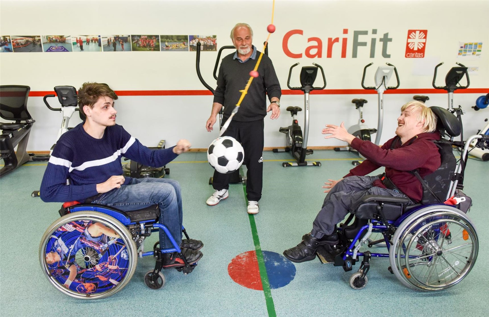Zwei Männer im Rollstuhl spielen Ball. Ein Betreuer steht zwischen ihnen.