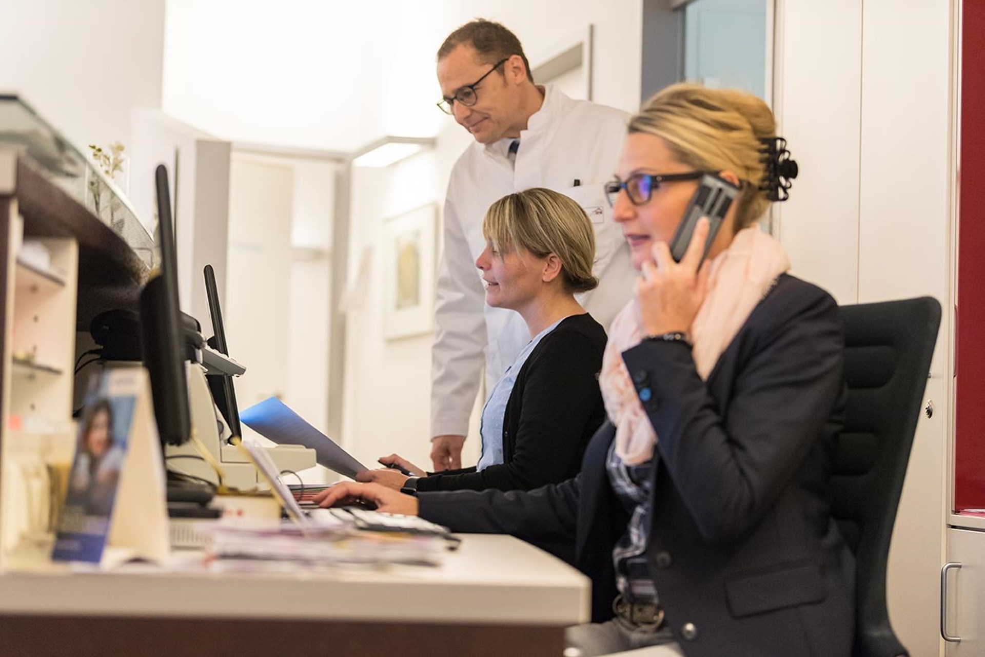Zwei Mitarbeiterinnen eines Krankenhauses sitzen an ihren Schreibtischen mit Telefon und Computer