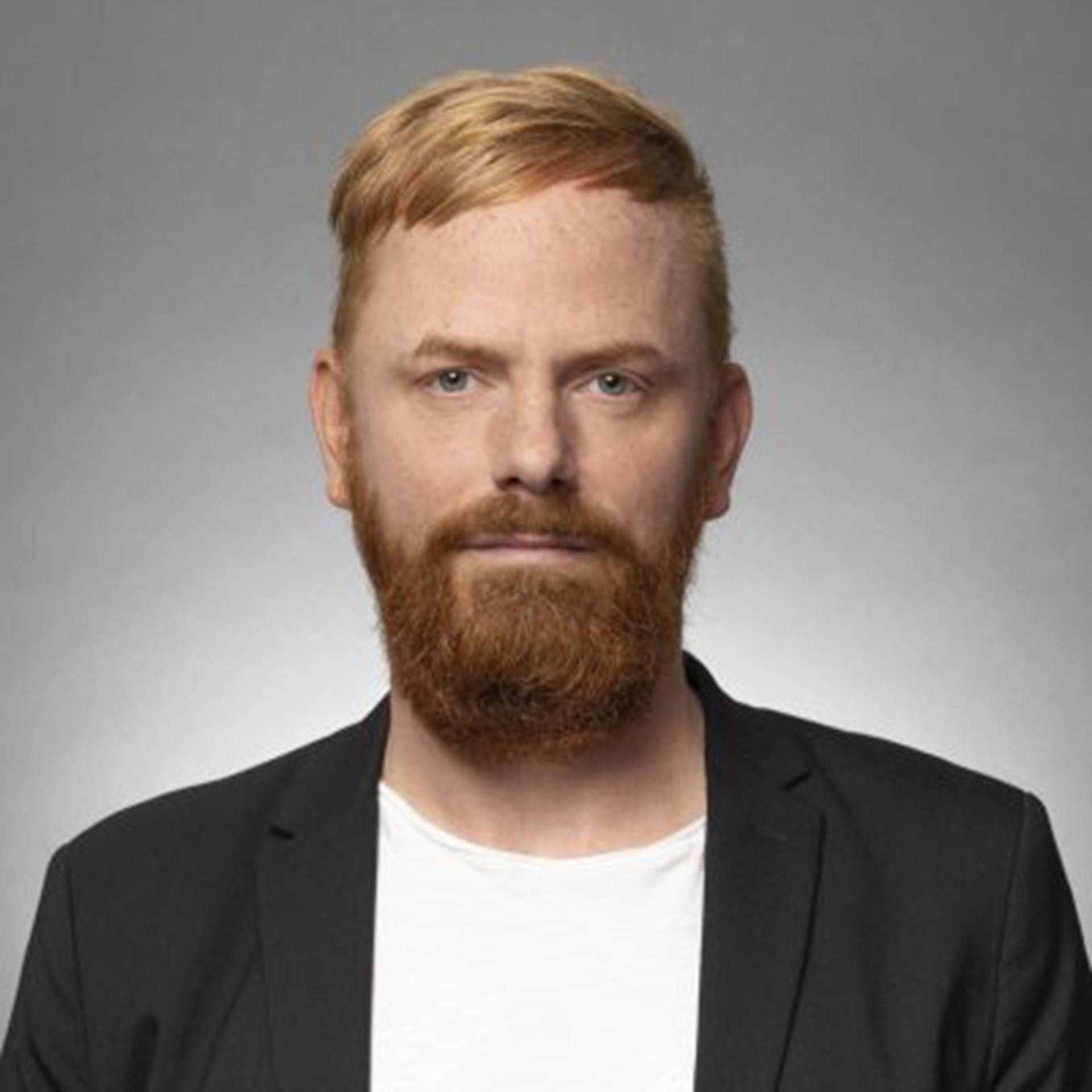 Mann mit roten Haaren und Vollbart, der ein weißes T-Shirt und eine schwarze Anzugjacke trägt